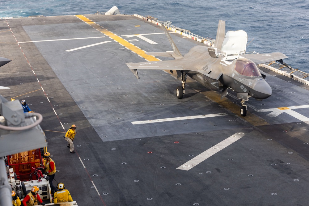 An F-35B Lightning II stealth fighter jet is positioned on the flight deck of an amphibious assault ship, likely a U.S. Navy vessel. Deck crew members in yellow and red safety gear are visible on the port side of the flight deck. The ocean is visible in the background, indicating the ship is underway at sea.