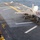 USS Boxer Conducts Flight Operations An F-35B Lightning II stealth fighter jet is positioned on the flight deck of an amphibious assault ship, likely a U.S. Navy vessel. Deck crew members in yellow and red safety gear are visible on the port side of the flight deck. The ocean is visible in the background, indicating the ship is underway at sea.