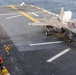 An F-35B Lightning II stealth fighter jet is positioned on the flight deck of an amphibious assault ship, likely a U.S. Navy vessel. Deck crew members in yellow and red safety gear are visible on the port side of the flight deck. The ocean is visible in the background, indicating the ship is underway at sea.