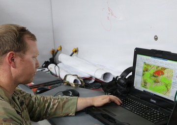 A U.S. Army soldier wearing 82nd Airborne Division insignia is working at a desk on a ruggedized Getac laptop displaying what appears to be a topographic or geospatial mapping application with color-coded terrain overlays. The setting appears to be a military command post or operations center, indicated by the whiteboard, rolled maps, and tactical equipment visible in the background. The soldier is in full OCP (Operational Camouflage Pattern) uniform with an American flag patch and Airborne tab.