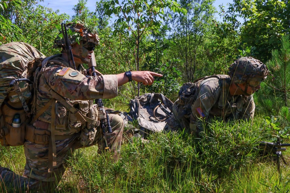 Two U.S. Army soldiers in full combat gear are operating in a wooded, vegetated area. The standing soldier is pointing and directing the other soldier who is in a low crouching position. Both wear OCP (Operational Camouflage Pattern) uniforms with American flag patches, suggesting a training exercise or field operation.