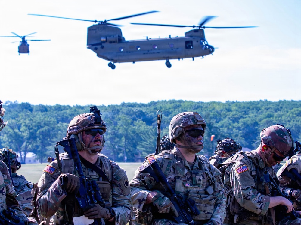 A group of fully equipped U.S. Army soldiers in MultiCam camouflage uniforms are staged in a field while two CH-47 Chinook helicopters operate overhead. The soldiers are wearing combat gear including helmets, body armor, face camouflage paint, and carrying rifles, suggesting a training exercise or air assault operation. The setting appears to be a military training area with open fields and tree lines in the background.