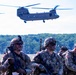 A group of fully equipped U.S. Army soldiers in MultiCam camouflage uniforms are staged in a field while two CH-47 Chinook helicopters operate overhead. The soldiers are wearing combat gear including helmets, body armor, face camouflage paint, and carrying rifles, suggesting a training exercise or air assault operation. The setting appears to be a military training area with open fields and tree lines in the background.