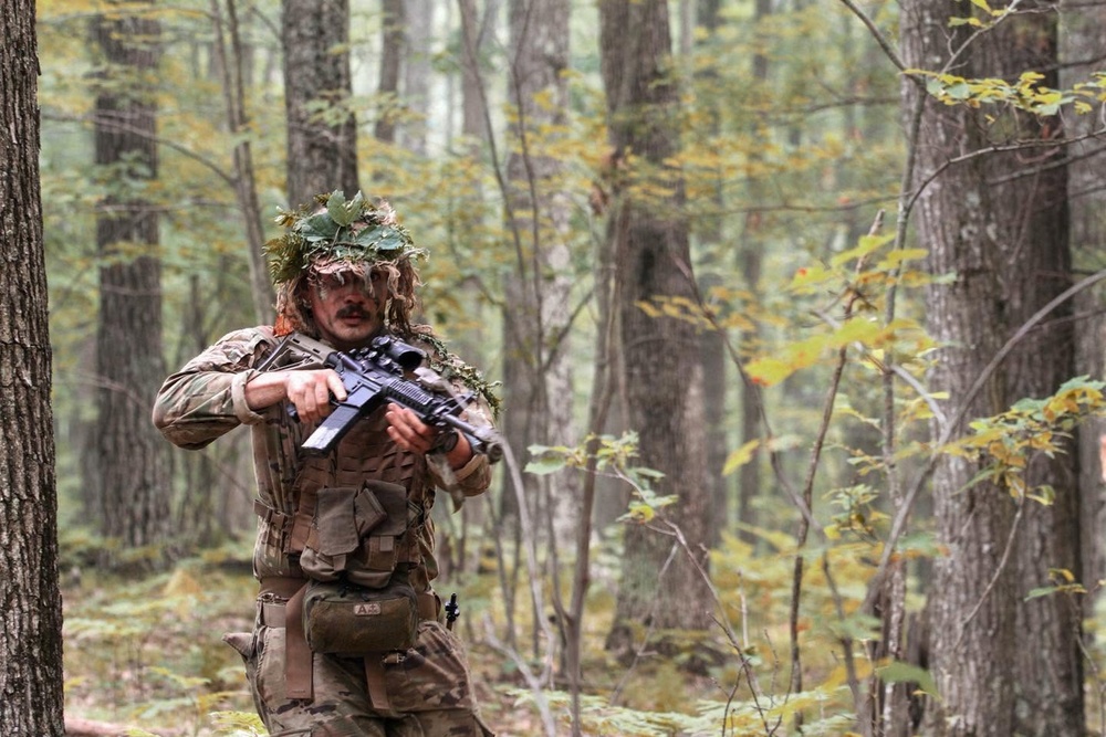 A soldier or tactical operator in full camouflage gear is moving through a deciduous forest, holding a rifle in a ready position. The individual is wearing a ghillie-style head covering with natural foliage attached for concealment, along with a tactical vest and multicam-pattern uniform. The setting appears to be a training exercise or field operation during autumn, based on the changing leaf colors.
