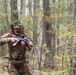 A soldier or tactical operator in full camouflage gear is moving through a deciduous forest, holding a rifle in a ready position. The individual is wearing a ghillie-style head covering with natural foliage attached for concealment, along with a tactical vest and multicam-pattern uniform. The setting appears to be a training exercise or field operation during autumn, based on the changing leaf colors.