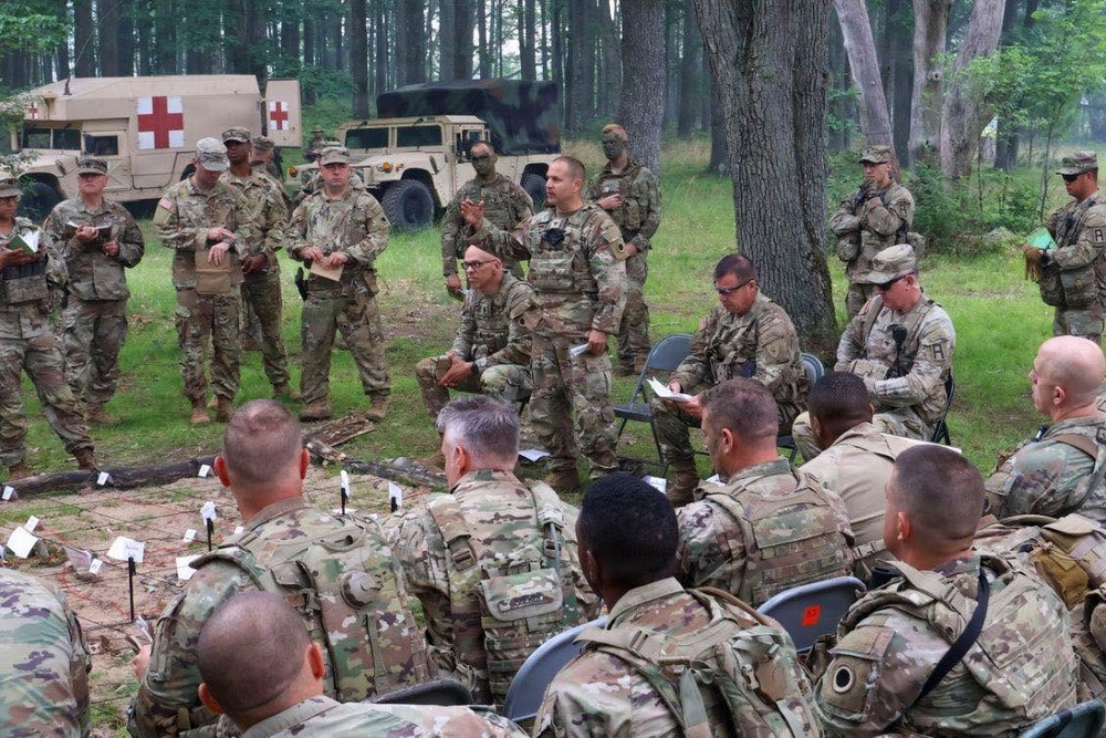 A group of U.S. Army soldiers in Operational Camouflage Pattern (OCP) uniforms are gathered in a wooded outdoor area for what appears to be a military briefing or after-action review. A central figure is standing and addressing the group, while others are seated in chairs or standing around the perimeter taking notes. Two military vehicles, including a medical HMMWV marked with a Red Cross, are visible in the background.