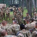 A group of U.S. Army soldiers in Operational Camouflage Pattern (OCP) uniforms are gathered in a wooded outdoor area for what appears to be a military briefing or after-action review. A central figure is standing and addressing the group, while others are seated in chairs or standing around the perimeter taking notes. Two military vehicles, including a medical HMMWV marked with a Red Cross, are visible in the background.