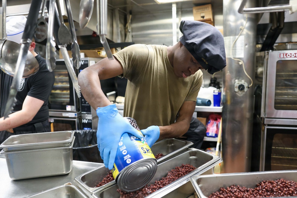 A food service worker wearing blue nitrile gloves and a gray hair net is pouring canned beans into a stainless steel hotel pan in a commercial kitchen. A second worker is visible in the background near additional kitchen equipment. The setting appears to be a large institutional or military galley kitchen.