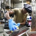 A food service worker wearing blue nitrile gloves and a gray hair net is pouring canned beans into a stainless steel hotel pan in a commercial kitchen. A second worker is visible in the background near additional kitchen equipment. The setting appears to be a large institutional or military galley kitchen.