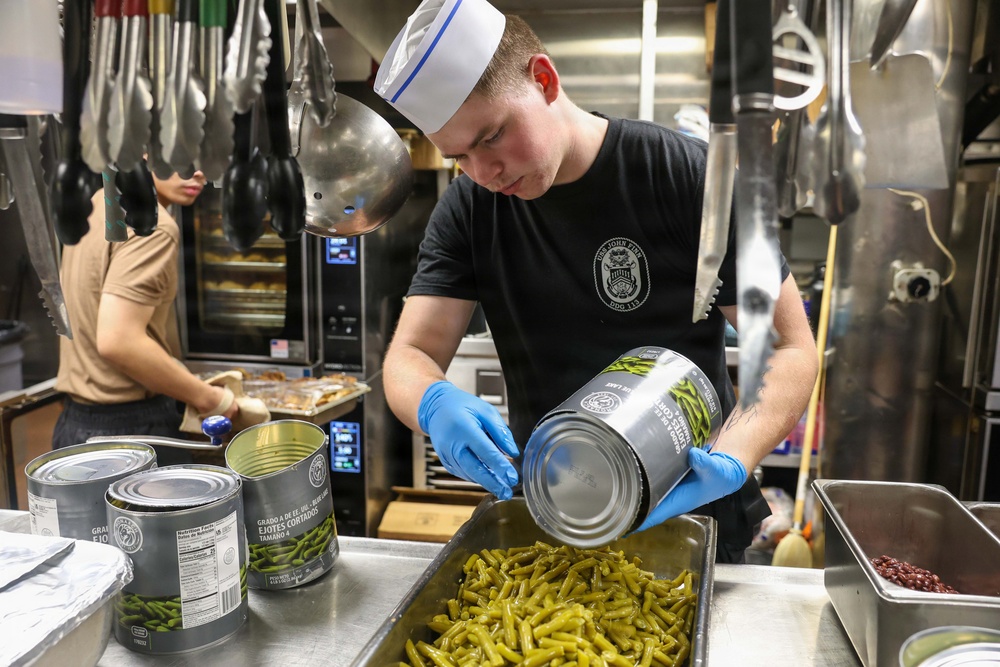 A young cook wearing a white chef's hat and black t-shirt with a military ship insignia is preparing food in a professional kitchen, pouring canned green beans into a large metal tray. Multiple large institutional-sized cans of cut green beans are visible on the stainless steel counter. A second worker in a tan shirt is visible in the background working at another station.