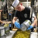 A young cook wearing a white chef's hat and black t-shirt with a military ship insignia is preparing food in a professional kitchen, pouring canned green beans into a large metal tray. Multiple large institutional-sized cans of cut green beans are visible on the stainless steel counter. A second worker in a tan shirt is visible in the background working at another station.