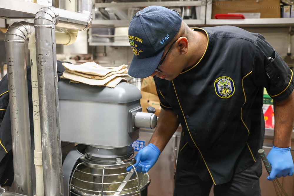 A Navy sailor assigned to USS John Finn (DDG-113) is working in a ship's galley kitchen, operating or adjusting a large commercial stand mixer. The individual is wearing a black uniform with gold trim bearing the USS John Finn patch, a matching ball cap, glasses, and blue nitrile food-handling gloves. The setting appears to be a compact, industrial naval kitchen environment.