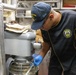 A Navy sailor assigned to USS John Finn (DDG-113) is working in a ship's galley kitchen, operating or adjusting a large commercial stand mixer. The individual is wearing a black uniform with gold trim bearing the USS John Finn patch, a matching ball cap, glasses, and blue nitrile food-handling gloves. The setting appears to be a compact, industrial naval kitchen environment.