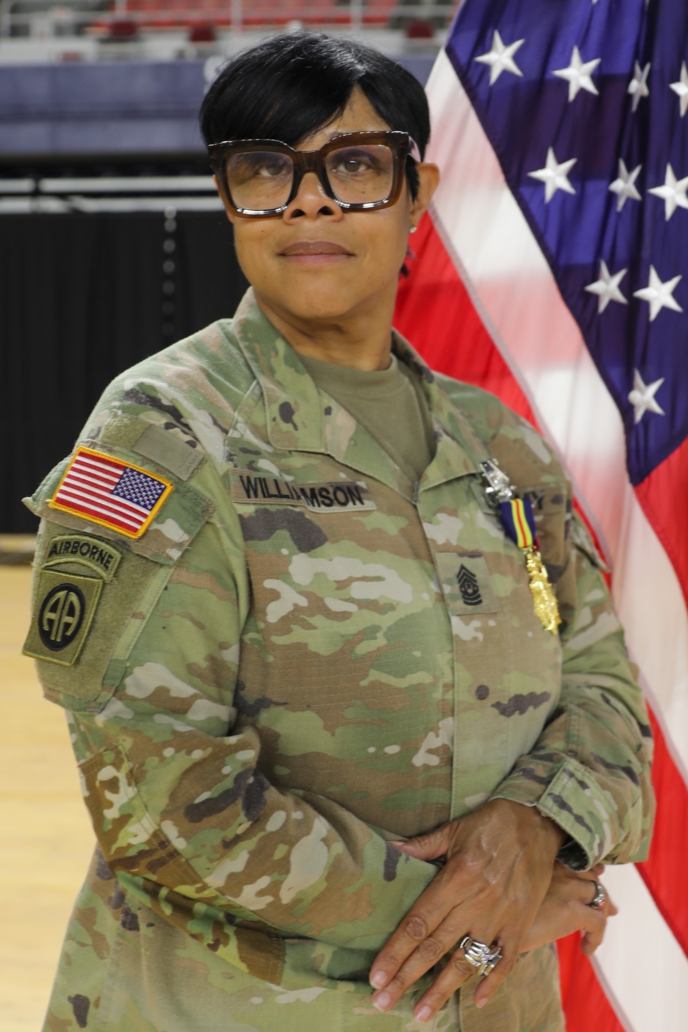 A U.S. Army soldier named Williamson, wearing Operational Camouflage Pattern (OCP) uniform, poses in front of an American flag. She displays the rank insignia of Sergeant Major (SGM) or Command Sergeant Major (CSM) on her chest, along with the 82nd Airborne Division patch and Airborne tab on her left shoulder. She is wearing large-framed glasses and appears to be at an official military ceremony or formal event.