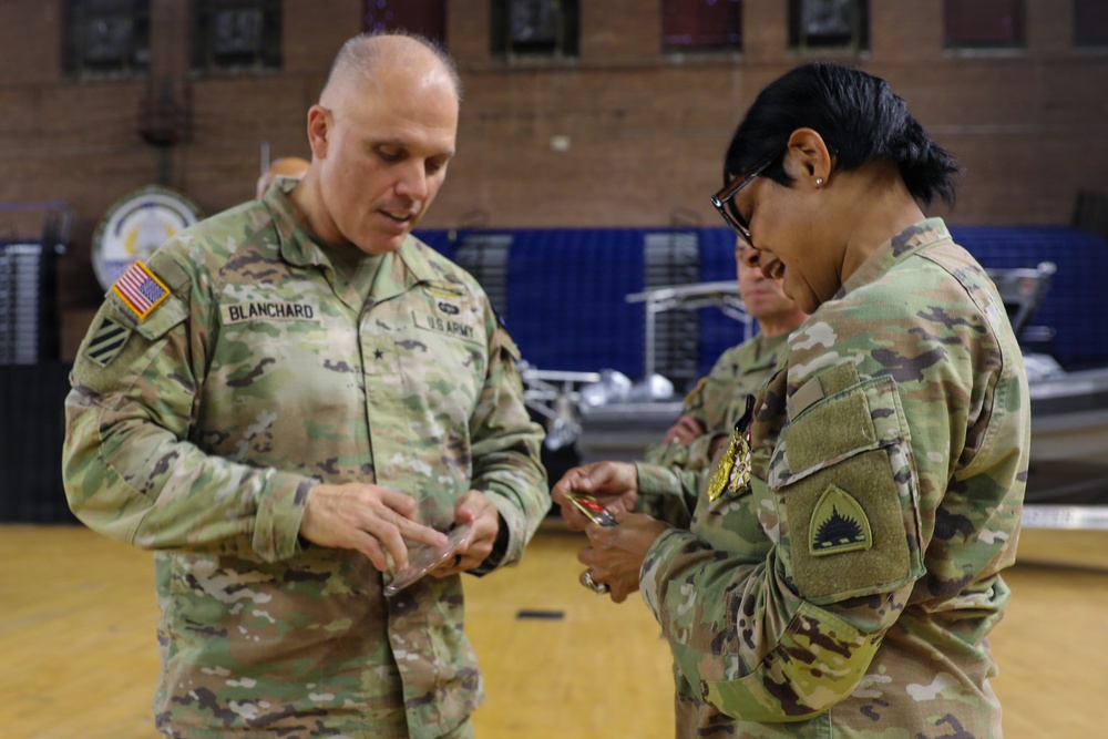 Two U.S. Army soldiers in OCP (Operational Camouflage Pattern) uniforms are engaged in what appears to be an award or decoration ceremony inside a gymnasium or armory. The soldier on the left, identified by name tape as 'BLANCHARD,' appears to be presenting or examining an item with the soldier on the right, who is wearing a medal. A third soldier is partially visible in the background.