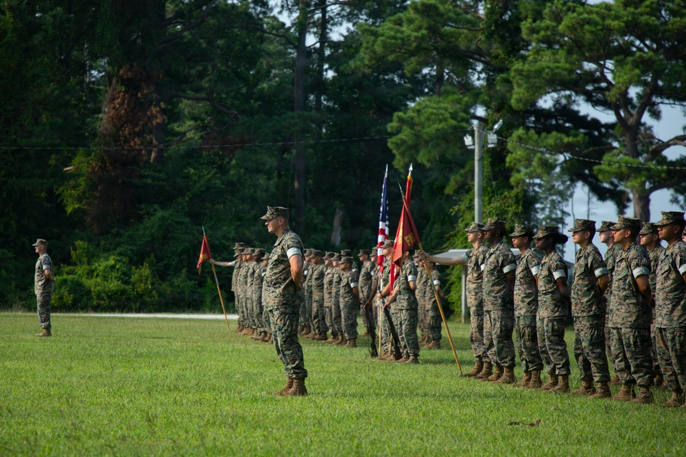 A formal military parade or ceremony is taking place on a grass field, featuring personnel in U.S. Marine Corps MARPAT camouflage uniforms standing in organized formation. Multiple flags are visible including the American flag and what appear to be unit guidons. A senior figure stands facing the formation, suggesting a command review, change of command, or similar ceremonial event.