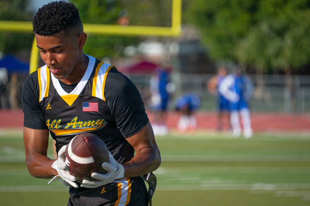 A male athlete wearing an 'All Army' black and gold football jersey with an American flag patch holds a football on a sports field. The foreground subject is in sharp focus while the background shows blurred figures in blue and white uniforms near a goalpost. The setting appears to be a military all-star football event held outdoors during daylight hours.