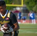 A male athlete wearing an 'All Army' black and gold football jersey with an American flag patch holds a football on a sports field. The foreground subject is in sharp focus while the background shows blurred figures in blue and white uniforms near a goalpost. The setting appears to be a military all-star football event held outdoors during daylight hours.