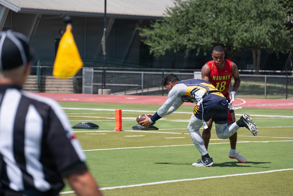 A flag football game is in progress on an outdoor turf field under bright sunlight. A player in a navy and gold uniform is reaching down to pick up or place the football near an orange pylon at what appears to be the goal line, while a player wearing a red 'Marines #18' jersey defends. A referee in a black and white striped shirt is visible in the foreground, with a yellow penalty flag on a pole nearby.