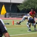 A flag football game is in progress on an outdoor turf field under bright sunlight. A player in a navy and gold uniform is reaching down to pick up or place the football near an orange pylon at what appears to be the goal line, while a player wearing a red 'Marines #18' jersey defends. A referee in a black and white striped shirt is visible in the foreground, with a yellow penalty flag on a pole nearby.
