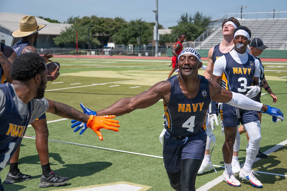 A flag football game featuring players wearing Navy-branded uniforms on an outdoor turf field. The central player, wearing number 4, is celebrating with an outstretched arm reaching toward a teammate wearing orange and blue gloves. Several other Navy teammates are visible in the background along with what appears to be a referee.