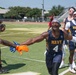 A flag football game featuring players wearing Navy-branded uniforms on an outdoor turf field. The central player, wearing number 4, is celebrating with an outstretched arm reaching toward a teammate wearing orange and blue gloves. Several other Navy teammates are visible in the background along with what appears to be a referee.
