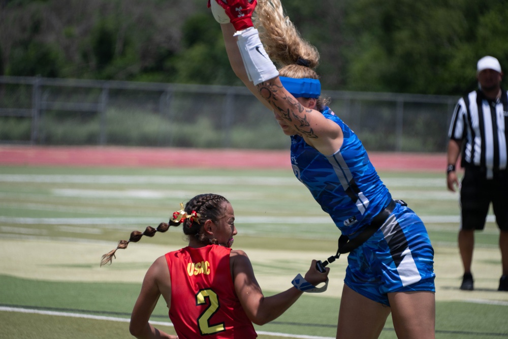 A flag football game is in progress on a turf field, showing two female players in a competitive moment where the blue-uniformed player leaps high to catch or deflect a pass while the red-uniformed player (number 2, OSAC) attempts to pull her flag. A referee in a black-and-white striped shirt observes the play from the background.