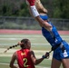 A flag football game is in progress on a turf field, showing two female players in a competitive moment where the blue-uniformed player leaps high to catch or deflect a pass while the red-uniformed player (number 2, OSAC) attempts to pull her flag. A referee in a black-and-white striped shirt observes the play from the background.