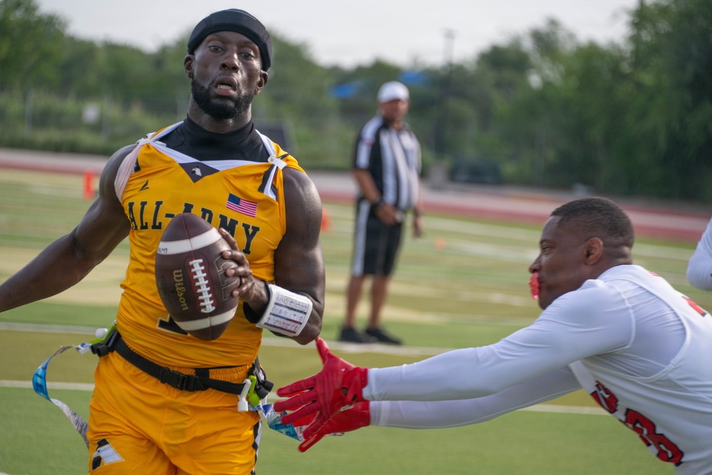 A flag football game is in progress, featuring a player in an 'All-Army' gold jersey carrying a Wilson football while an opposing player in white reaches to pull his flag. A referee in a black-and-white striped shirt observes the play in the background on a green turf field.
