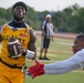 A flag football game is in progress, featuring a player in an 'All-Army' gold jersey carrying a Wilson football while an opposing player in white reaches to pull his flag. A referee in a black-and-white striped shirt observes the play in the background on a green turf field.