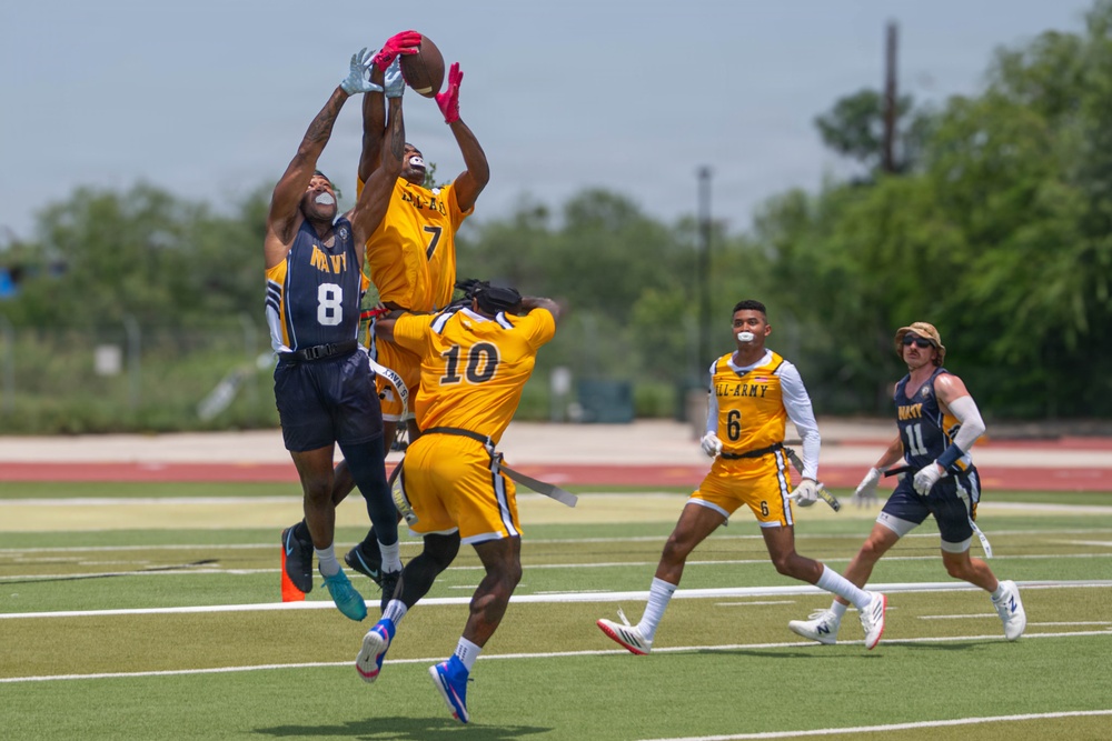 A flag football game is being played between a Navy team (dark blue uniforms) and an All-Army team (gold/yellow uniforms) on an artificial turf field. The focal action shows Navy player #8 and Army player #7 both leaping to contest a pass, with the football visible at the top of their reach. Additional players including Army #10, Army #6, and Navy #11 are visible in the background observing the play.
