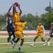 A flag football game is being played between a Navy team (dark blue uniforms) and an All-Army team (gold/yellow uniforms) on an artificial turf field. The focal action shows Navy player #8 and Army player #7 both leaping to contest a pass, with the football visible at the top of their reach. Additional players including Army #10, Army #6, and Navy #11 are visible in the background observing the play.
