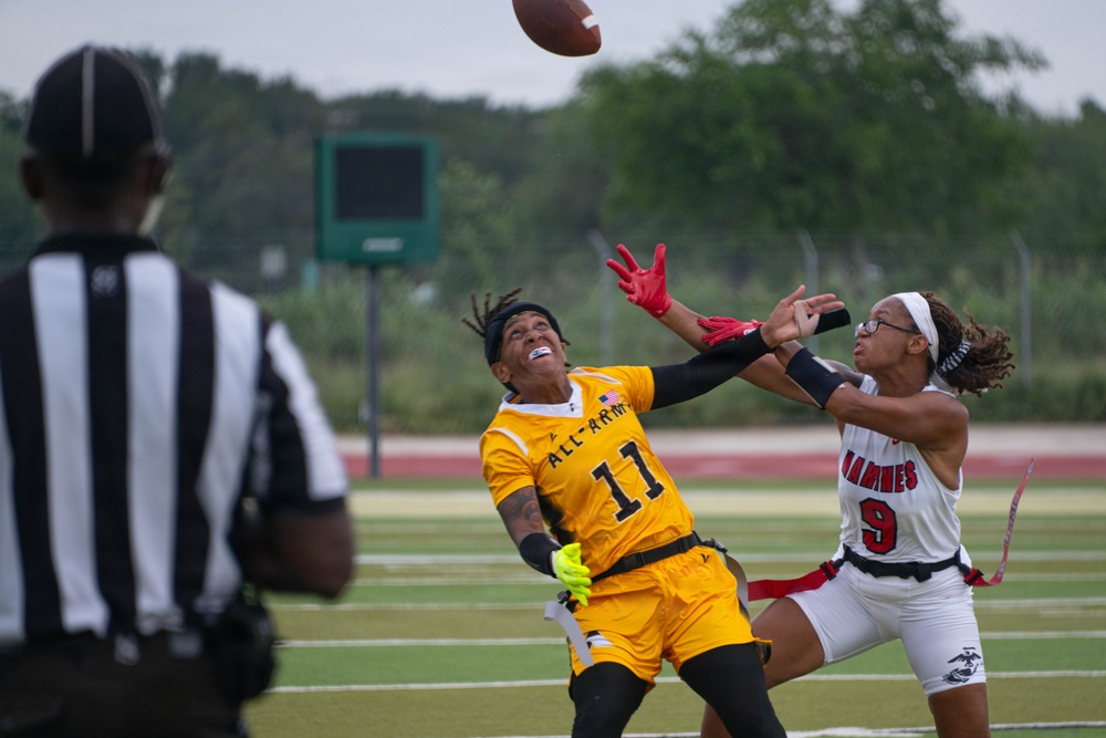 A flag football game between military branch teams — All-Army (gold uniforms) and Marines (white/red uniforms) — is captured mid-play as two female players contest a pass. Player #11 from All-Army and player #9 from the Marines are both reaching for a football in the air. A referee in a black-and-white striped shirt observes the play from the left foreground.
