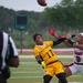 A flag football game between military branch teams — All-Army (gold uniforms) and Marines (white/red uniforms) — is captured mid-play as two female players contest a pass. Player #11 from All-Army and player #9 from the Marines are both reaching for a football in the air. A referee in a black-and-white striped shirt observes the play from the left foreground.