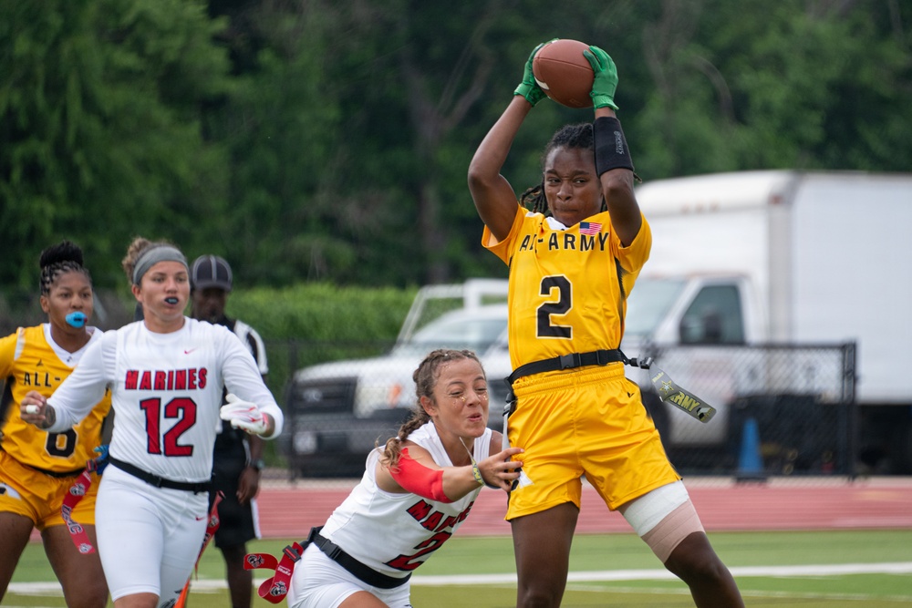 A women's flag football game between the All-Army and Marines military teams is in progress. A player wearing an All-Army gold jersey numbered 2 is catching or securing a football while a Marines defender in white attempts to interfere. Additional players from both teams are visible in the background on what appears to be a well-maintained athletic field.
