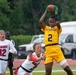 A women's flag football game between the All-Army and Marines military teams is in progress. A player wearing an All-Army gold jersey numbered 2 is catching or securing a football while a Marines defender in white attempts to interfere. Additional players from both teams are visible in the background on what appears to be a well-maintained athletic field.
