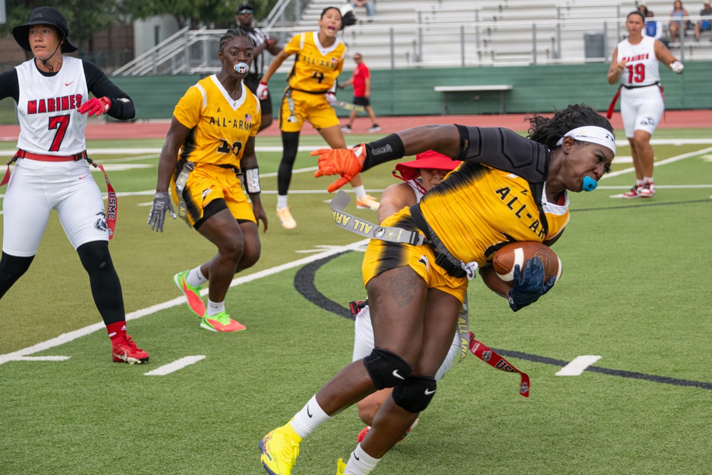 A women's flag football game between the All-Army team (gold/black uniforms) and the Marines team (white/red uniforms). A player wearing an All-Army jersey is making a dynamic catch or run with the football while being defended, leaning forward aggressively. The action is taking place on a green turf field with stadium bleachers visible in the background.