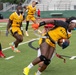 A women's flag football game between the All-Army team (gold/black uniforms) and the Marines team (white/red uniforms). A player wearing an All-Army jersey is making a dynamic catch or run with the football while being defended, leaning forward aggressively. The action is taking place on a green turf field with stadium bleachers visible in the background.