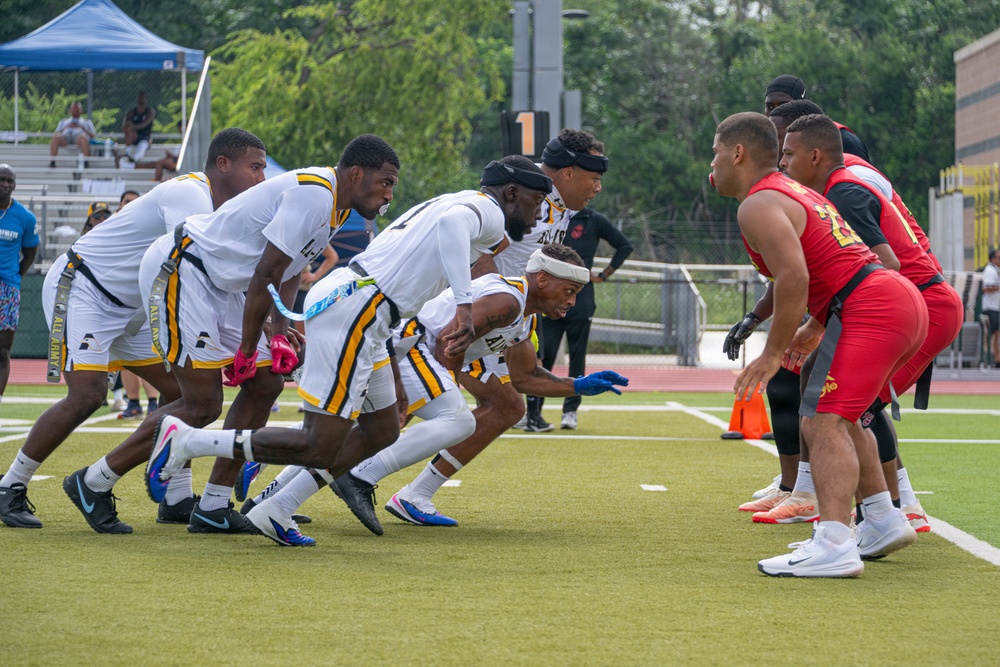 A flag football game is in progress on a green turf field, with two teams lined up at the line of scrimmage. The white-uniformed team with black and gold accents appears to be in an offensive formation, crouching and ready to snap the ball, while the red-uniformed team stands in a defensive position. Spectators and a blue canopy tent are visible in the background bleachers.