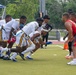 A flag football game is in progress on a green turf field, with two teams lined up at the line of scrimmage. The white-uniformed team with black and gold accents appears to be in an offensive formation, crouching and ready to snap the ball, while the red-uniformed team stands in a defensive position. Spectators and a blue canopy tent are visible in the background bleachers.