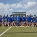 A large group of athletes and coaches pose for a team photo on an outdoor turf football field, many displaying medals around their necks or held up for the camera. The majority are wearing matching blue camouflage-patterned uniforms with 'Air Force' branding, suggesting this is a U.S. Air Force sports team. The setting appears to be a championship or tournament event, indicated by the medals and celebratory poses.