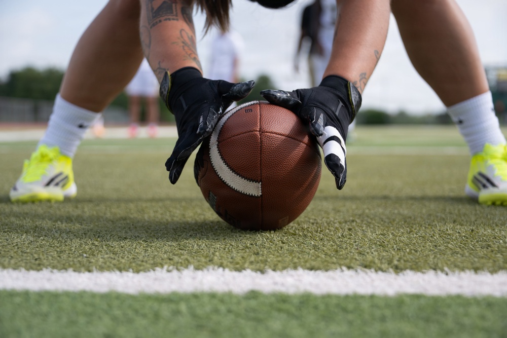A close-up shot of a player in a center stance position on an artificial turf football field, hands gripping a brown Wilson football on a yard line. The player is wearing black gloves, white socks, and neon yellow-green Adidas cleats, with visible tattoos on both forearms. Additional players are visible in the blurred background, suggesting a practice or game setting.