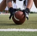 A close-up shot of a player in a center stance position on an artificial turf football field, hands gripping a brown Wilson football on a yard line. The player is wearing black gloves, white socks, and neon yellow-green Adidas cleats, with visible tattoos on both forearms. Additional players are visible in the blurred background, suggesting a practice or game setting.