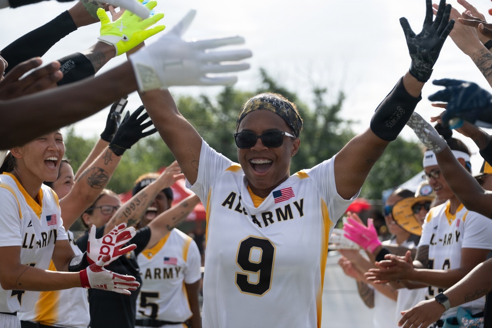 A female athlete wearing an All-Army jersey number 9 celebrates with arms raised while teammates form a celebratory tunnel around her. The scene captures a joyful post-game or pre-game moment with multiple players in white and gold All-Army uniforms clapping and cheering. The atmosphere is energetic and triumphant, suggesting a victory or significant achievement.