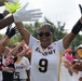 A female athlete wearing an All-Army jersey number 9 celebrates with arms raised while teammates form a celebratory tunnel around her. The scene captures a joyful post-game or pre-game moment with multiple players in white and gold All-Army uniforms clapping and cheering. The atmosphere is energetic and triumphant, suggesting a victory or significant achievement.