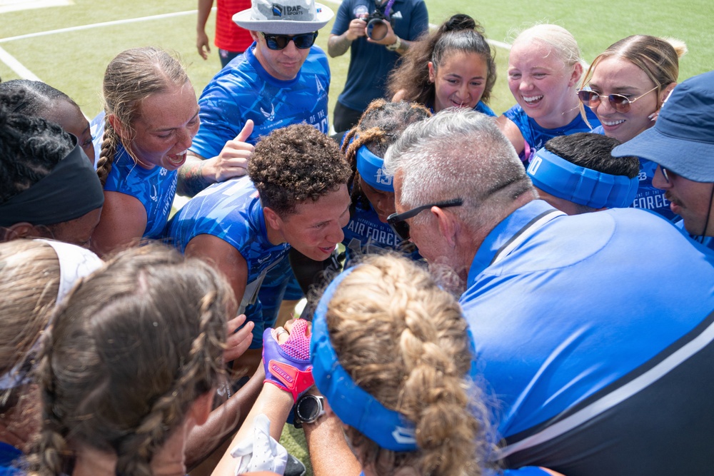 A group of athletes and coaches wearing U.S. Air Force branded blue jerseys are gathered in a celebratory team huddle on a sports field. The group appears to be in high spirits, with visible smiling, laughing, and enthusiastic expressions suggesting a post-game celebration or victory moment. The team appears to be a women's sports team, likely softball or flag football, based on visible equipment and uniforms.