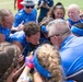 A group of athletes and coaches wearing U.S. Air Force branded blue jerseys are gathered in a celebratory team huddle on a sports field. The group appears to be in high spirits, with visible smiling, laughing, and enthusiastic expressions suggesting a post-game celebration or victory moment. The team appears to be a women's sports team, likely softball or flag football, based on visible equipment and uniforms.