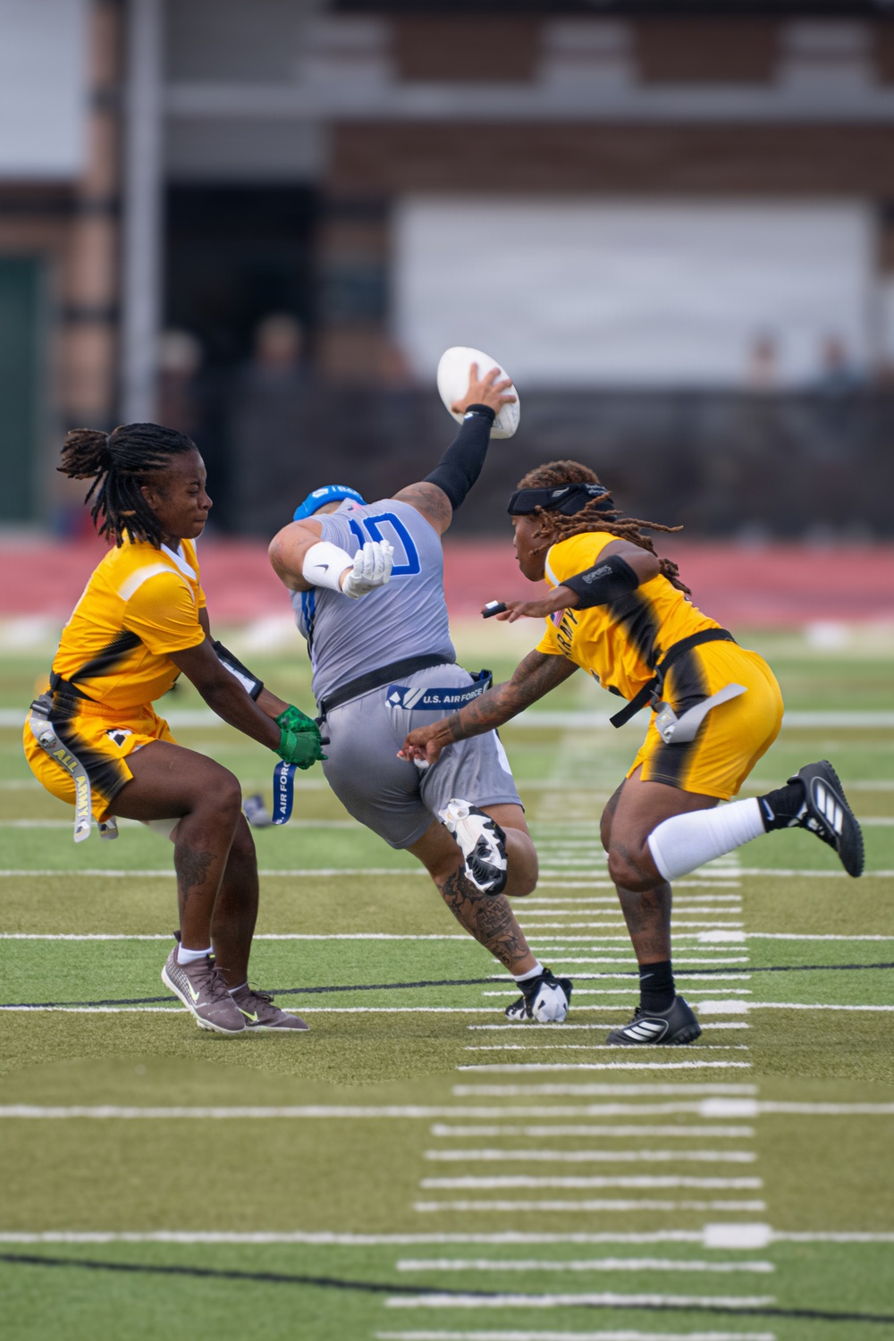 A flag football game is in progress on a green turf field, showing an intense play where a player in a gray U.S. Air Force uniform is releasing or throwing a football while being defended by two players in gold and black Army uniforms. The Air Force player is leaning dramatically backward while extending the ball upward, with both Army defenders closing in to pull his flags. The action is captured in sharp focus against a blurred stadium background.