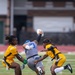 A flag football game is in progress on a green turf field, showing an intense play where a player in a gray U.S. Air Force uniform is releasing or throwing a football while being defended by two players in gold and black Army uniforms. The Air Force player is leaning dramatically backward while extending the ball upward, with both Army defenders closing in to pull his flags. The action is captured in sharp focus against a blurred stadium background.