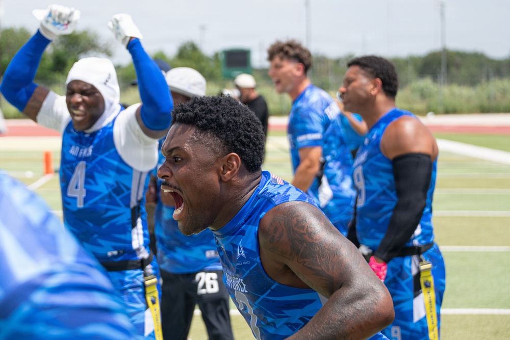A group of athletes wearing blue camouflage uniforms labeled 'Air Force' are celebrating intensely on a football sideline. The central figure is shouting with an expression of intense emotion, while teammates behind him raise their arms in celebration. The setting appears to be an outdoor athletic field during a flag football or similar competition.